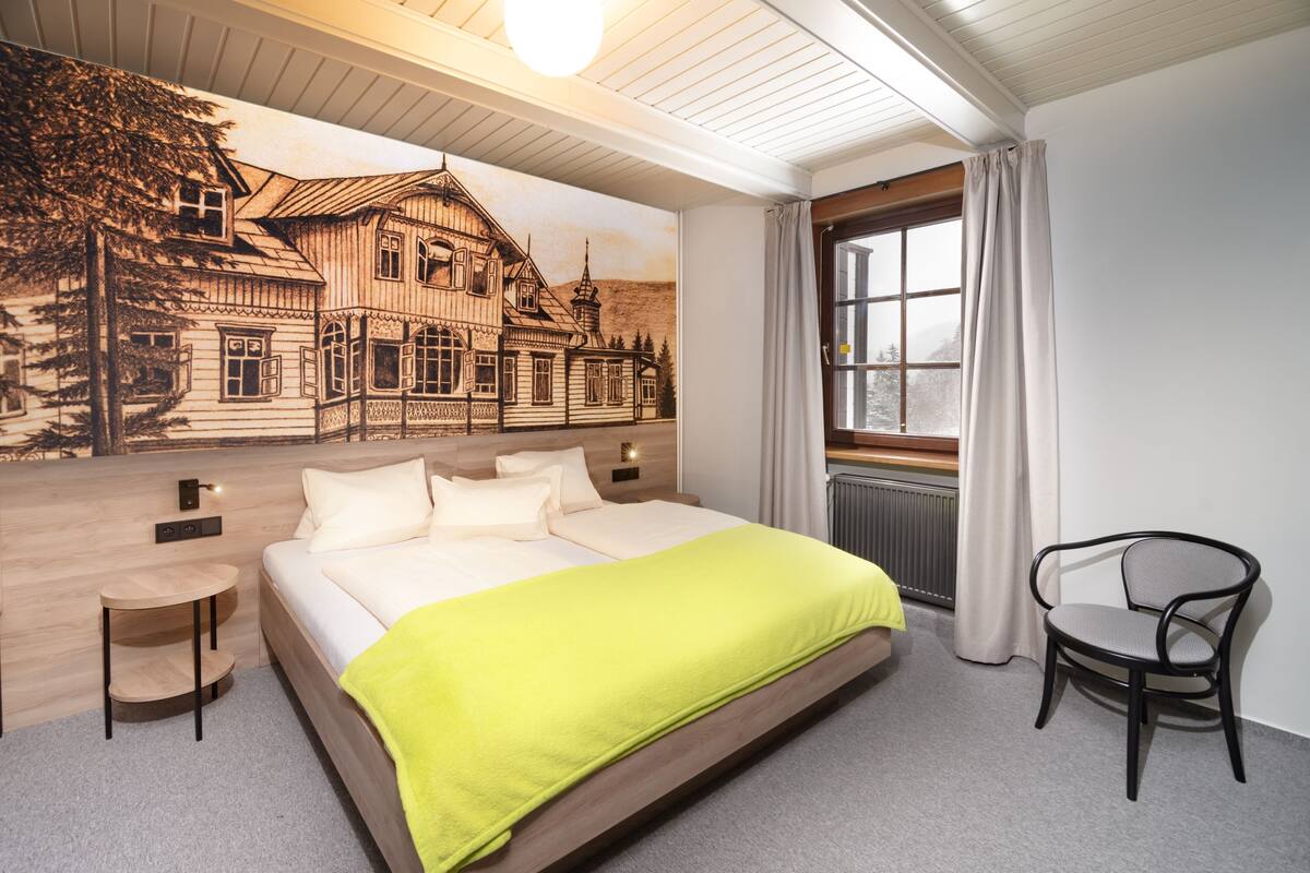 Ein modernes Schlafzimmer mit einem großen Bett, einem Nachttisch, einem Sessel, einem Fenster mit Vorhängen und einem Wandbild eines Hauses.