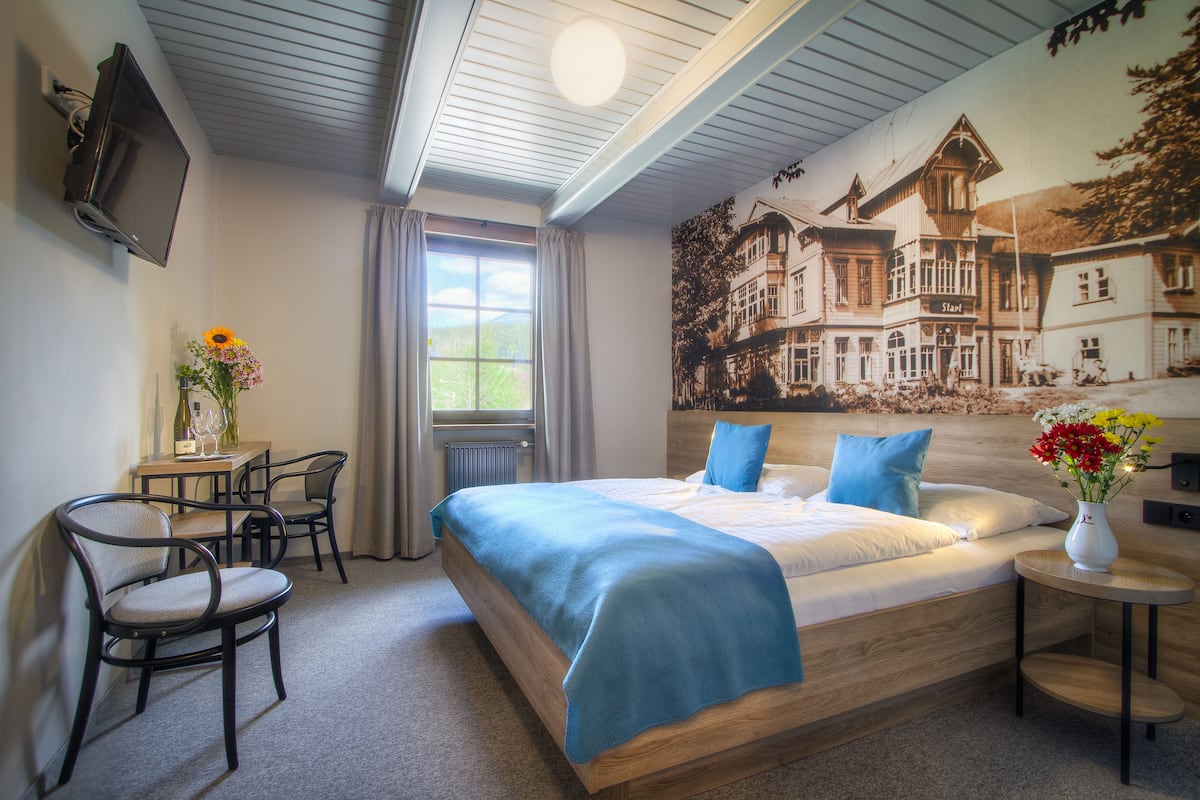 Ein Schlafzimmer mit einem Bett, einem Schreibtisch, zwei Stühlen, einer Vase mit Blumen und einem Bild an der Wand.