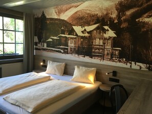 Quarto de hotel com uma cama grande, duas almofadas, uma mesa pequena e uma cadeira. Há uma janela com cortinas, um radiador e um mural na parede retratando uma paisagem nevada com edifícios.
