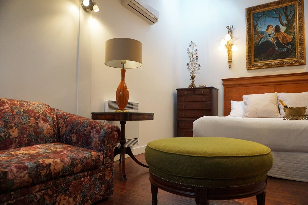 A hotel room with a floral sofa, a green ottoman, a wooden nightstand with a lamp, a dresser, and a bed with white linens.