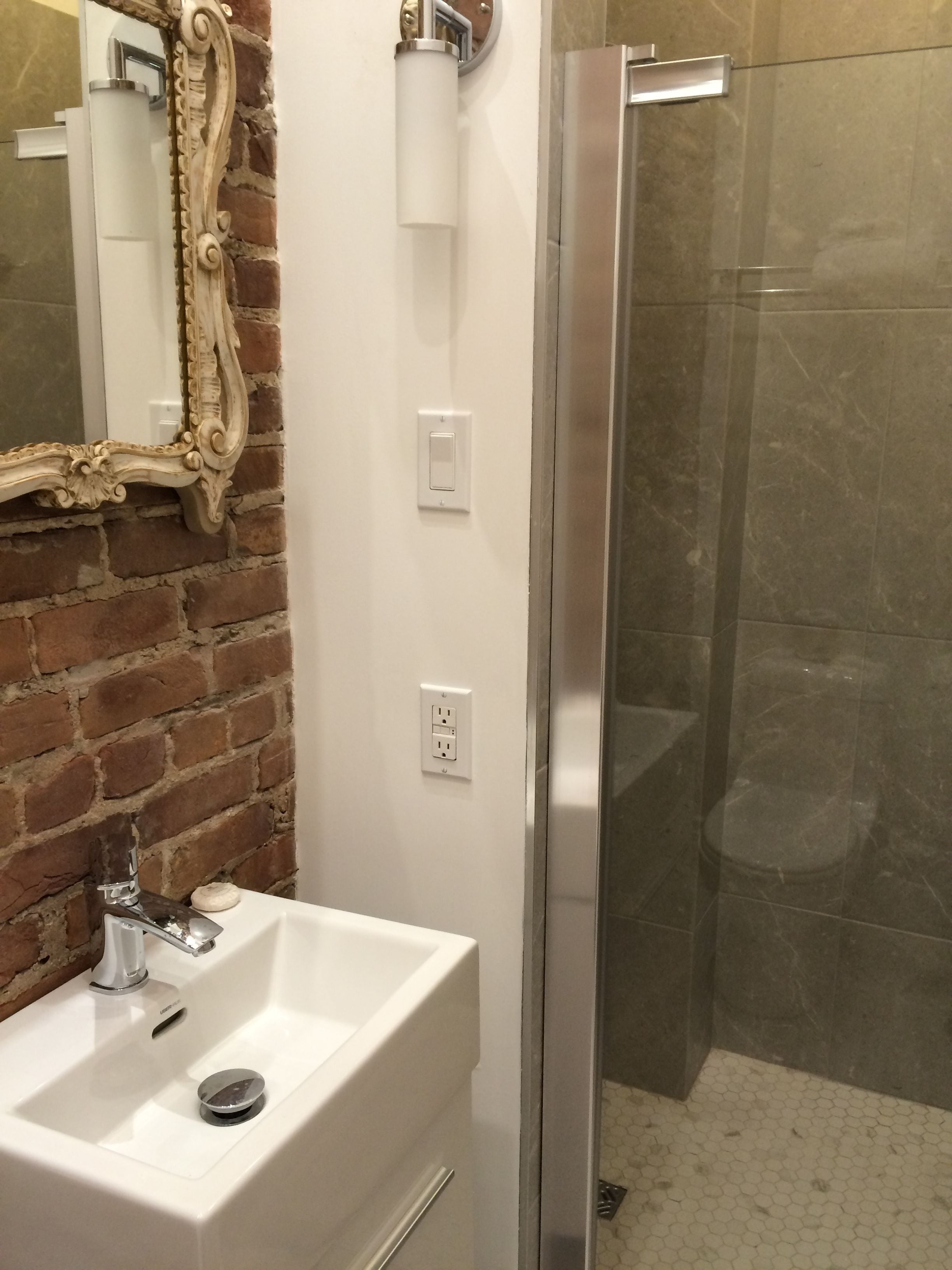 Un baño con un lavamanos blanco, un espejo con un marco decorativo, una ducha con puerta de vidrio y una pared de ladrillo.