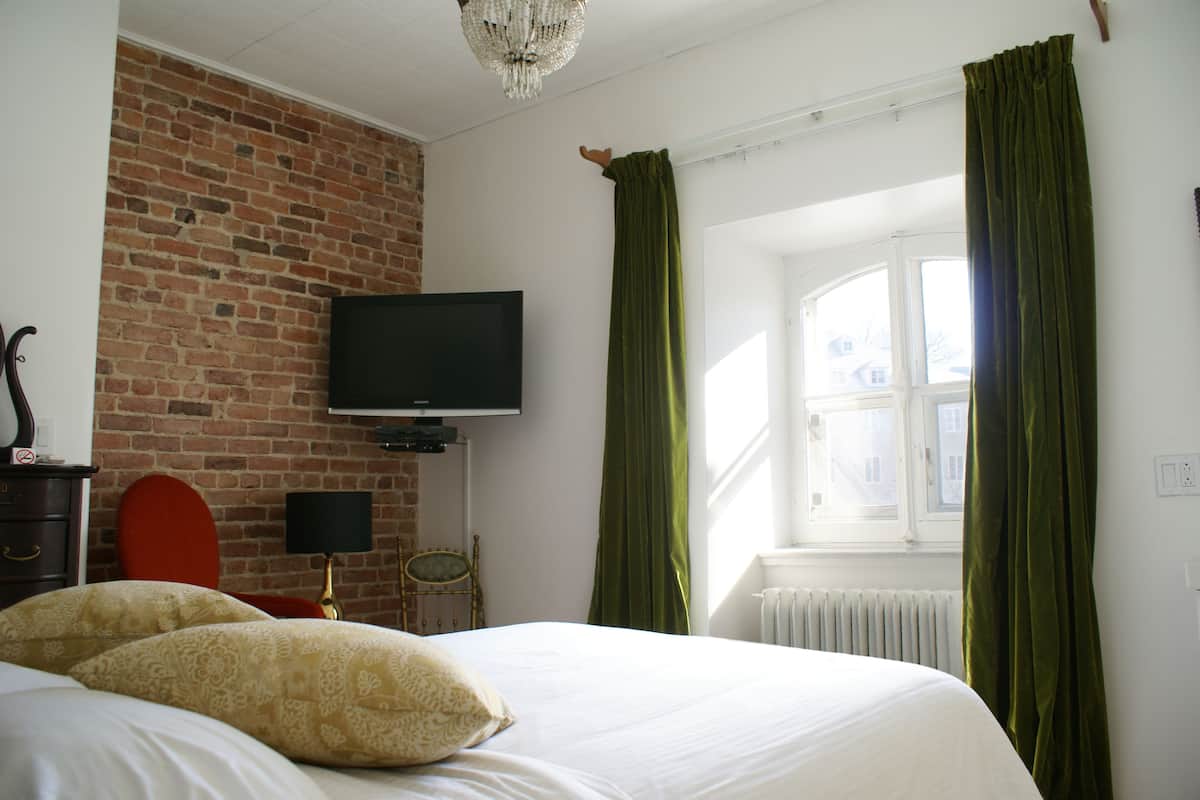 A hotel room with a brick wall, a flat-screen TV, a bed with white linens, green curtains, and a window with a view of buildings.