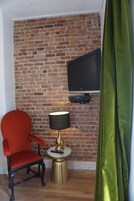 A room with a brick wall, a television mounted on it, a red chair, a small round table with a lamp, and a green curtain.