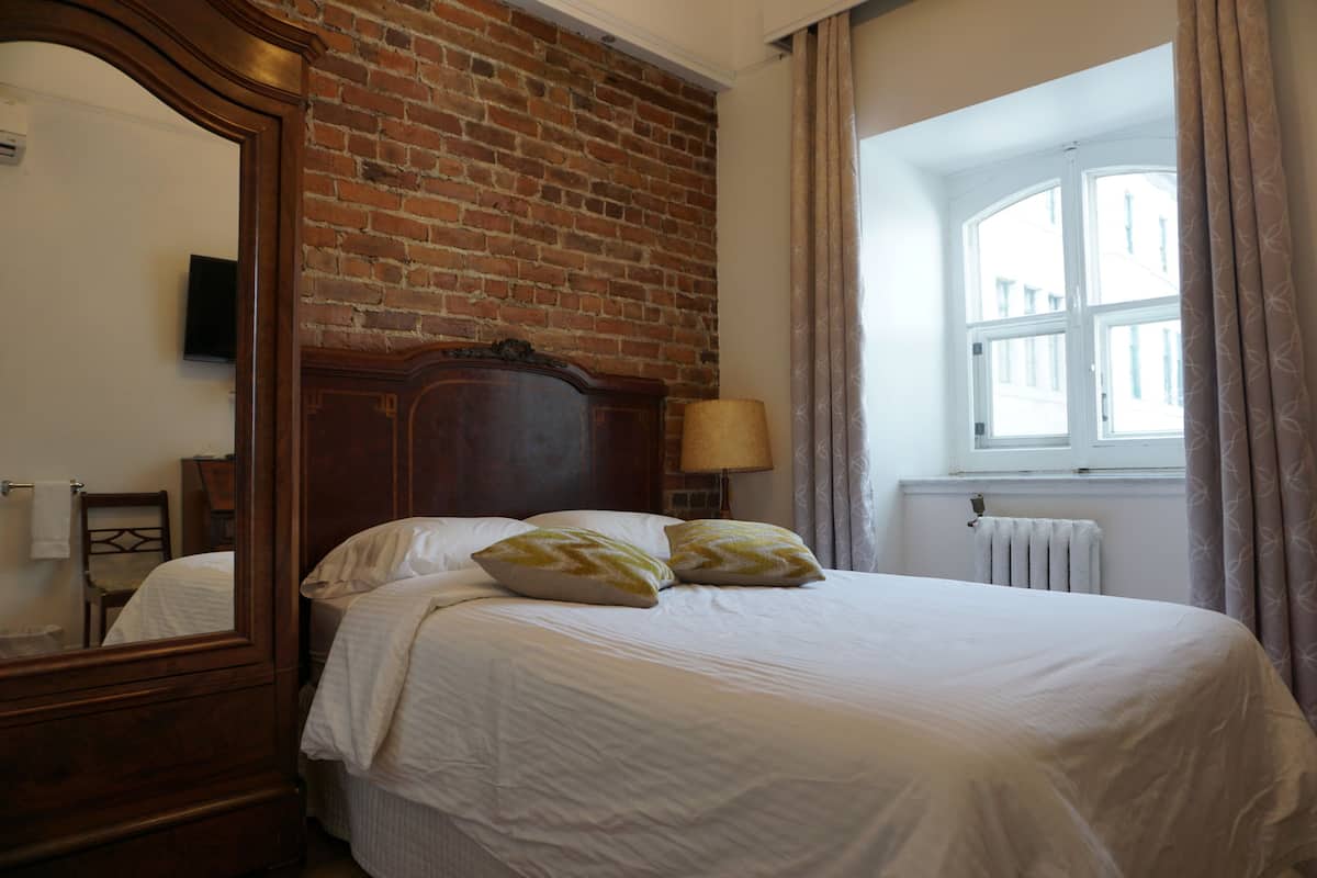 A hotel room with a brick wall, a large bed with white bedding, a wooden headboard, a mirror, a window with curtains, and a radiator.