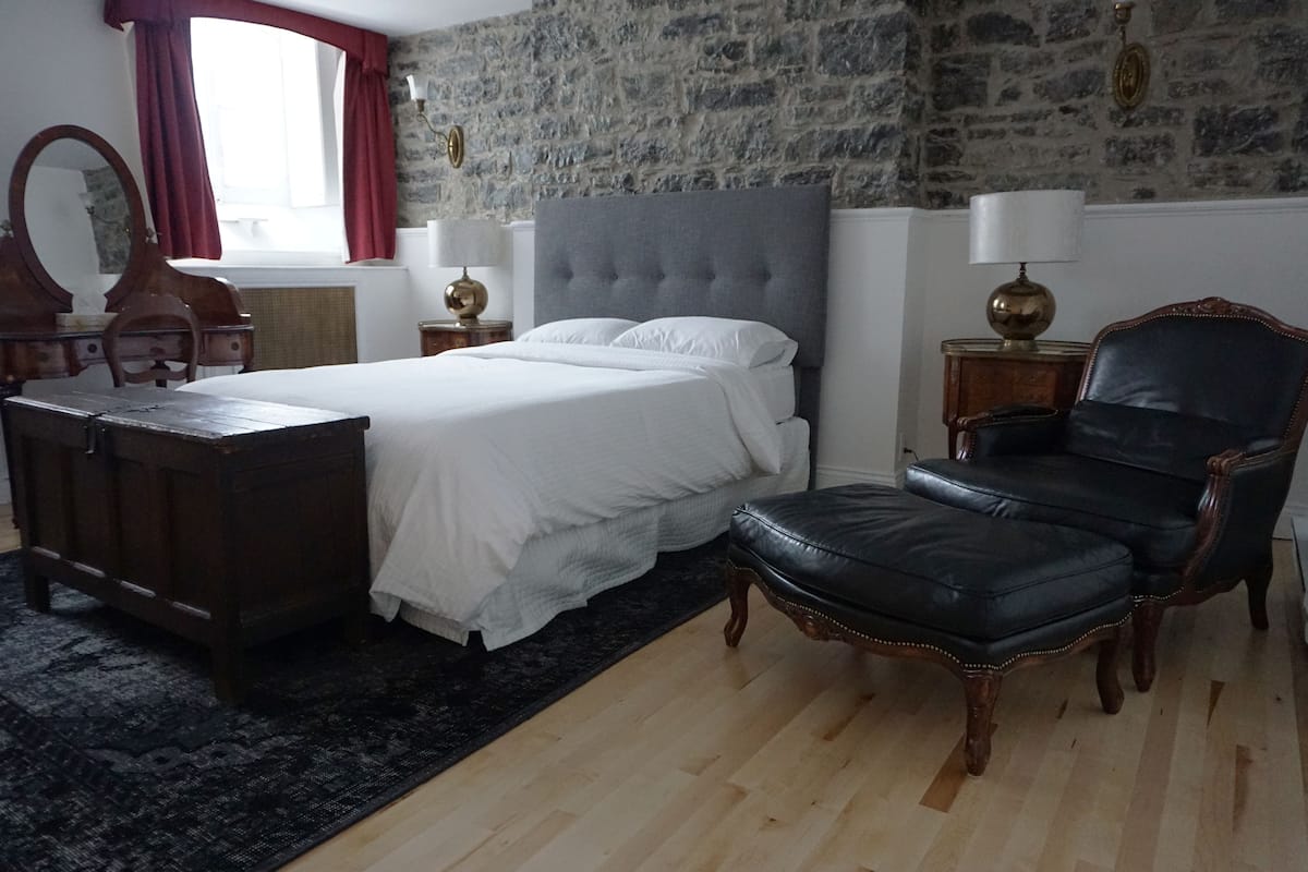 A bedroom with a large bed, a wooden dresser, a leather armchair, and a stone wall.