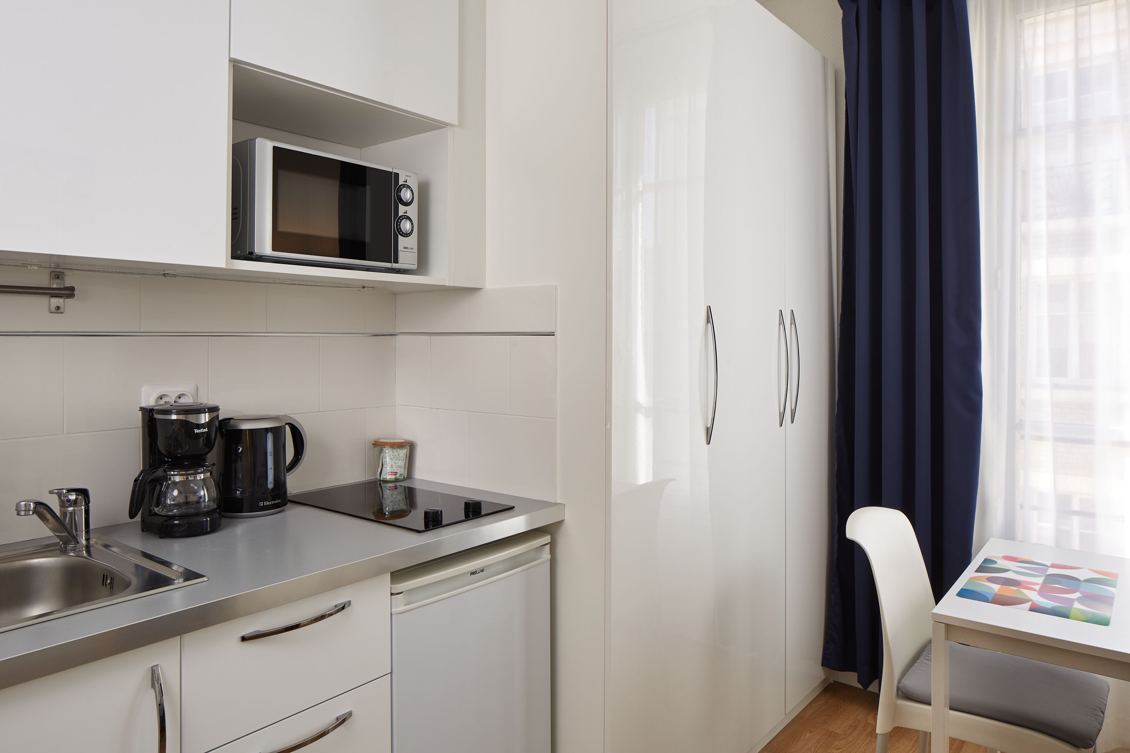 A modern kitchen with a microwave, coffee maker, and dishwasher. There is a white table with a colorful tablecloth and a white chair.
