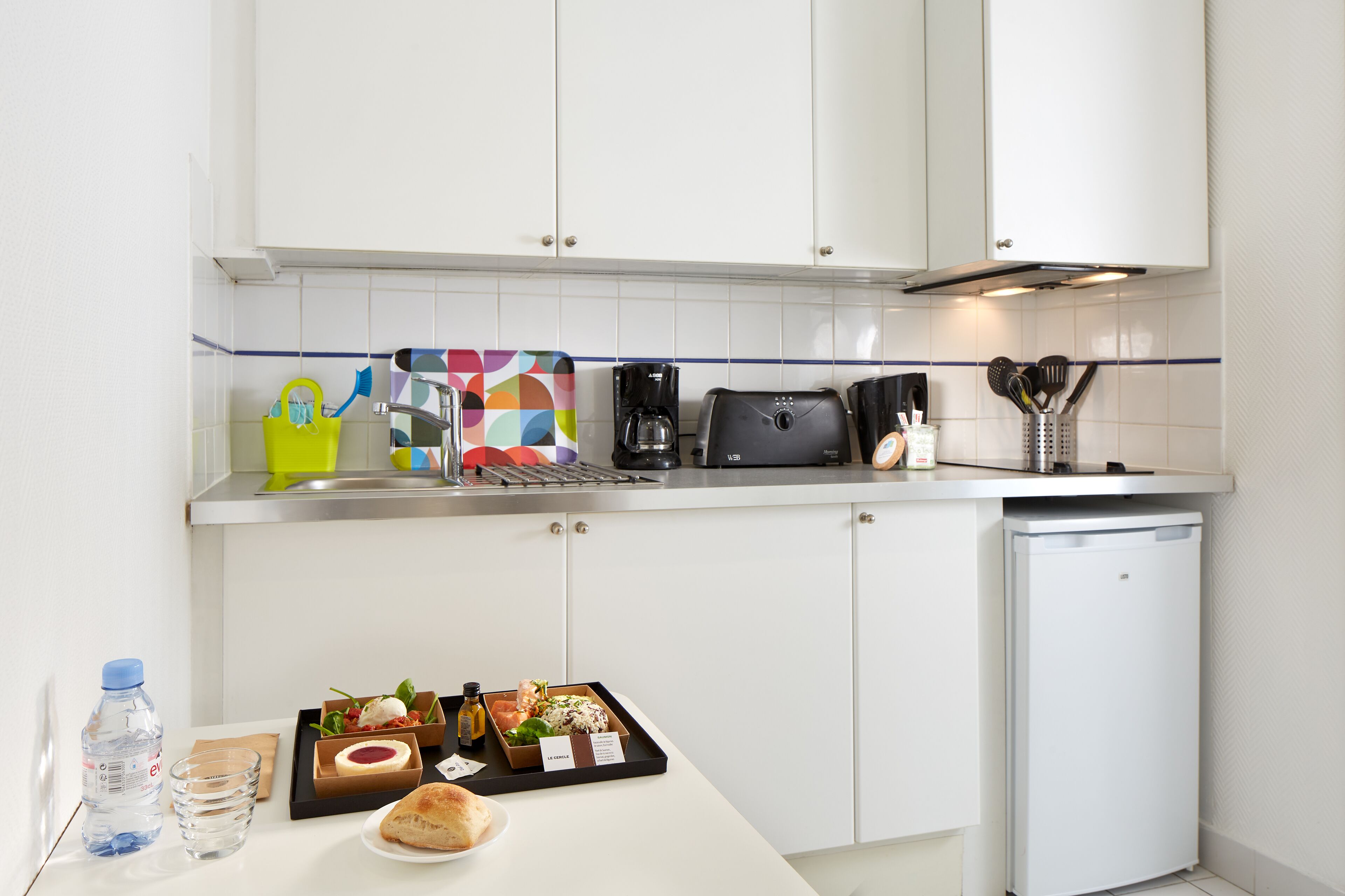 A modern kitchen with white cabinets, a stainless steel countertop, a coffee maker, a toaster, and a small refrigerator.