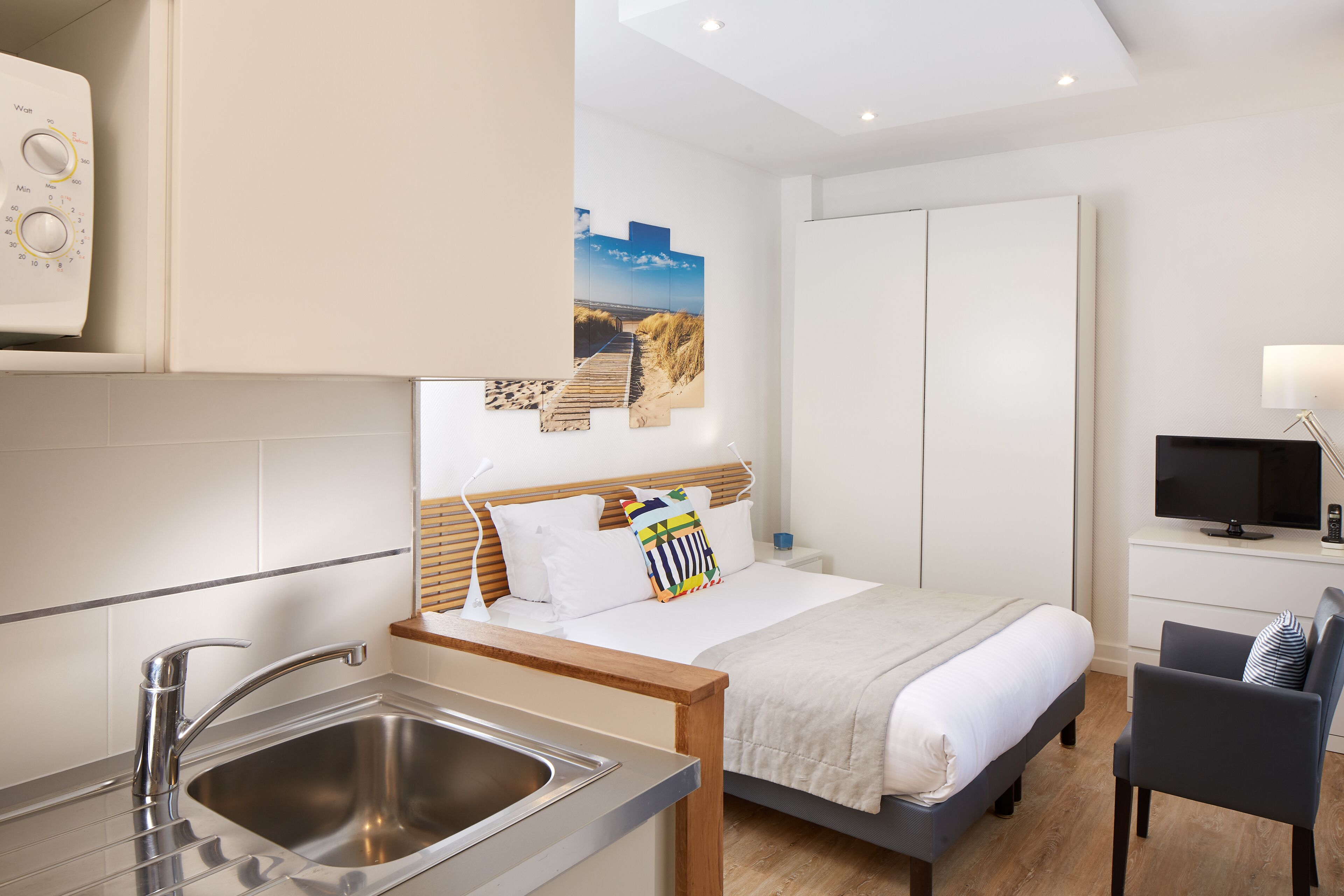 Een moderne hotelkamer met een houten bed, een ingebouwde televisie, een bureau met stoel, een wastafel en een schilderij van een strand.