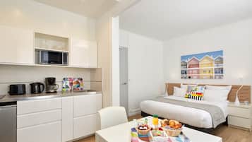 A modern hotel room with a kitchenette, a bed with a colorful pillow, a breakfast tray, and a painting of houses on the wall.