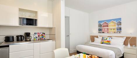 Una habitación de hotel moderna con kitchenette, una cama con un cojín colorido, una bandeja de desayuno y un cuadro de casas en la pared.