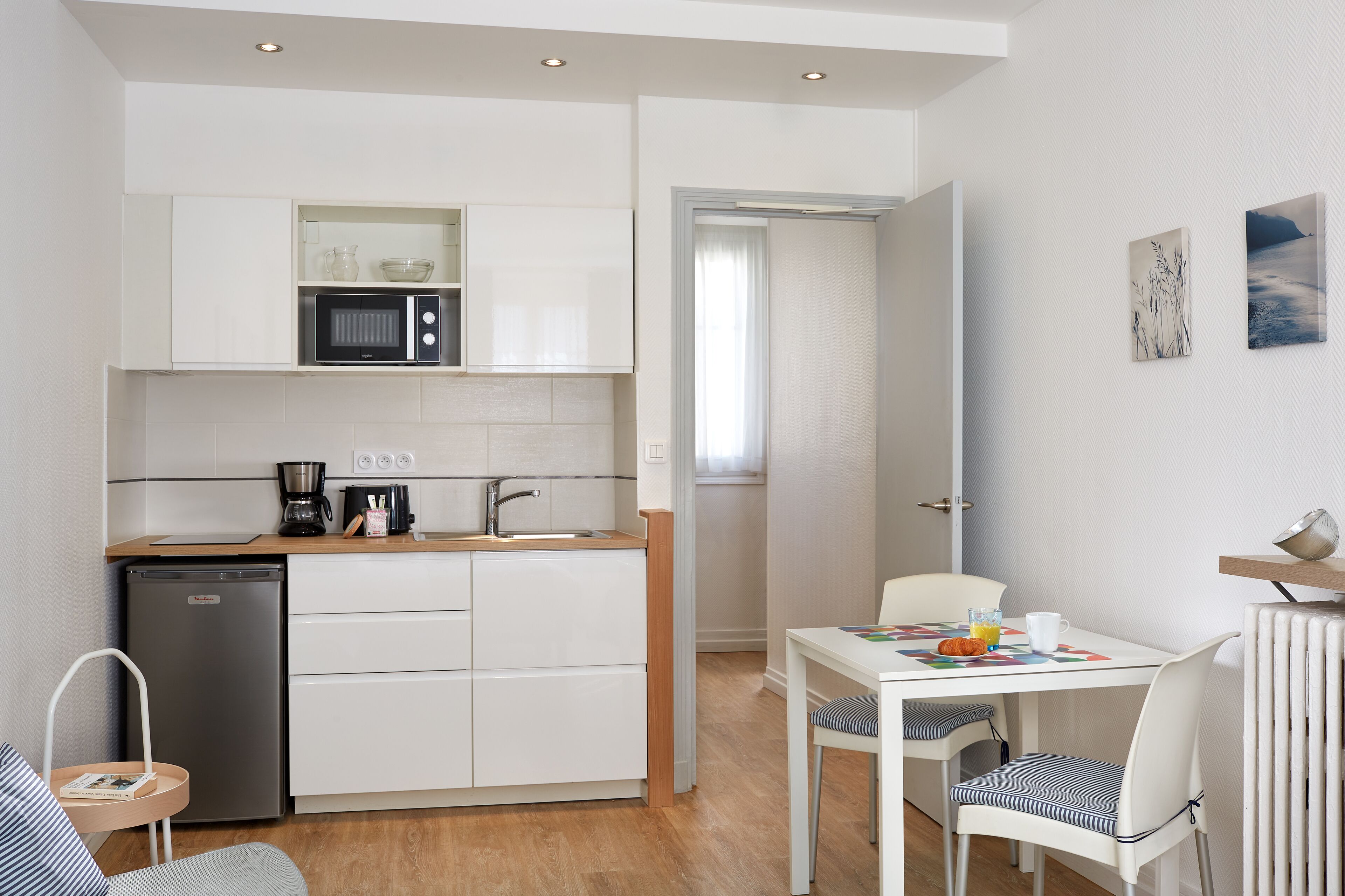 A compact kitchen with white cabinets, a microwave, and a coffee maker. There's a dining area with a table and chairs, and a small refrigerator.