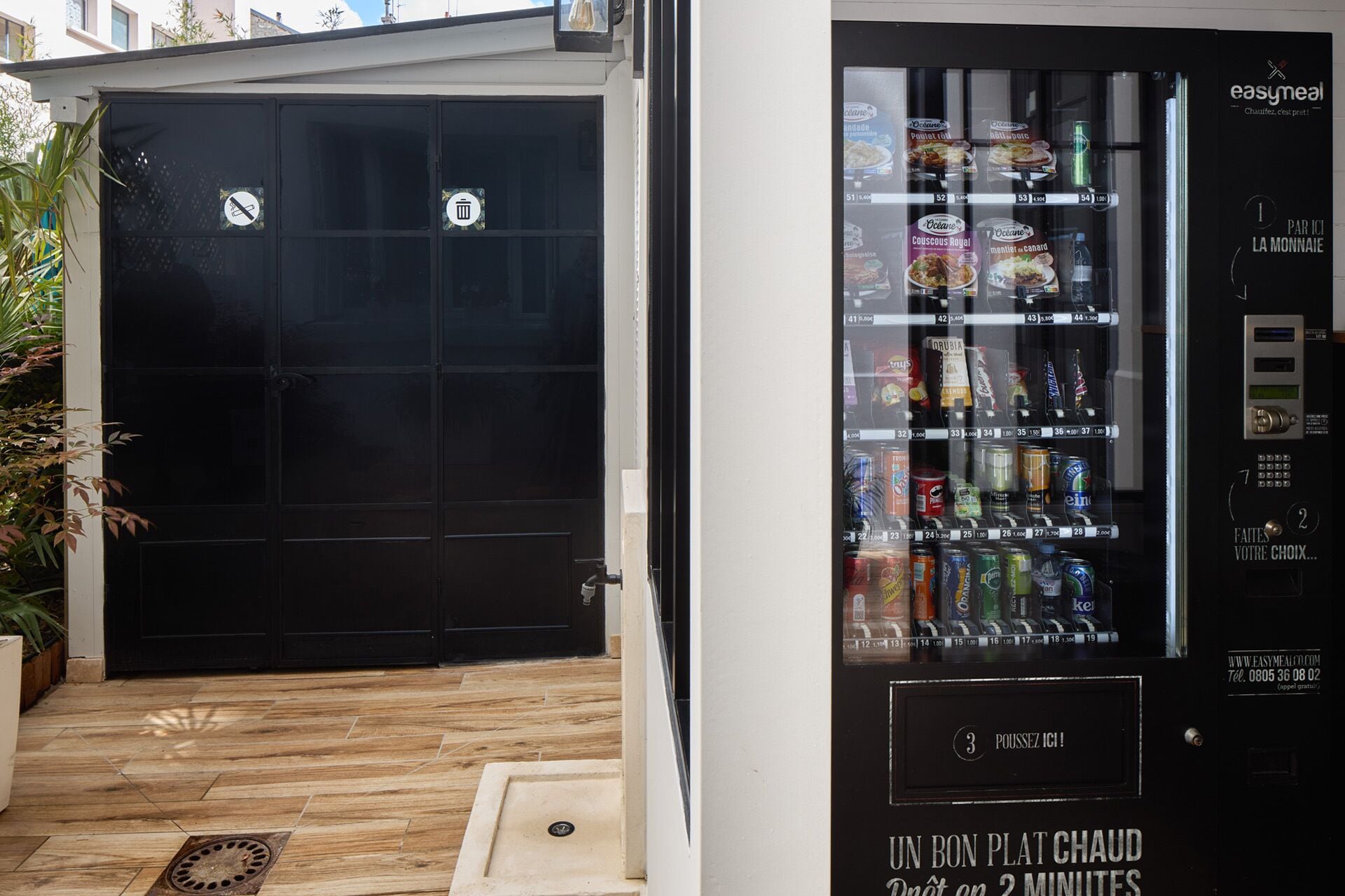 A vending machine with a variety of drinks and snacks, a black door with a trash bin, and a small step leading to the door.