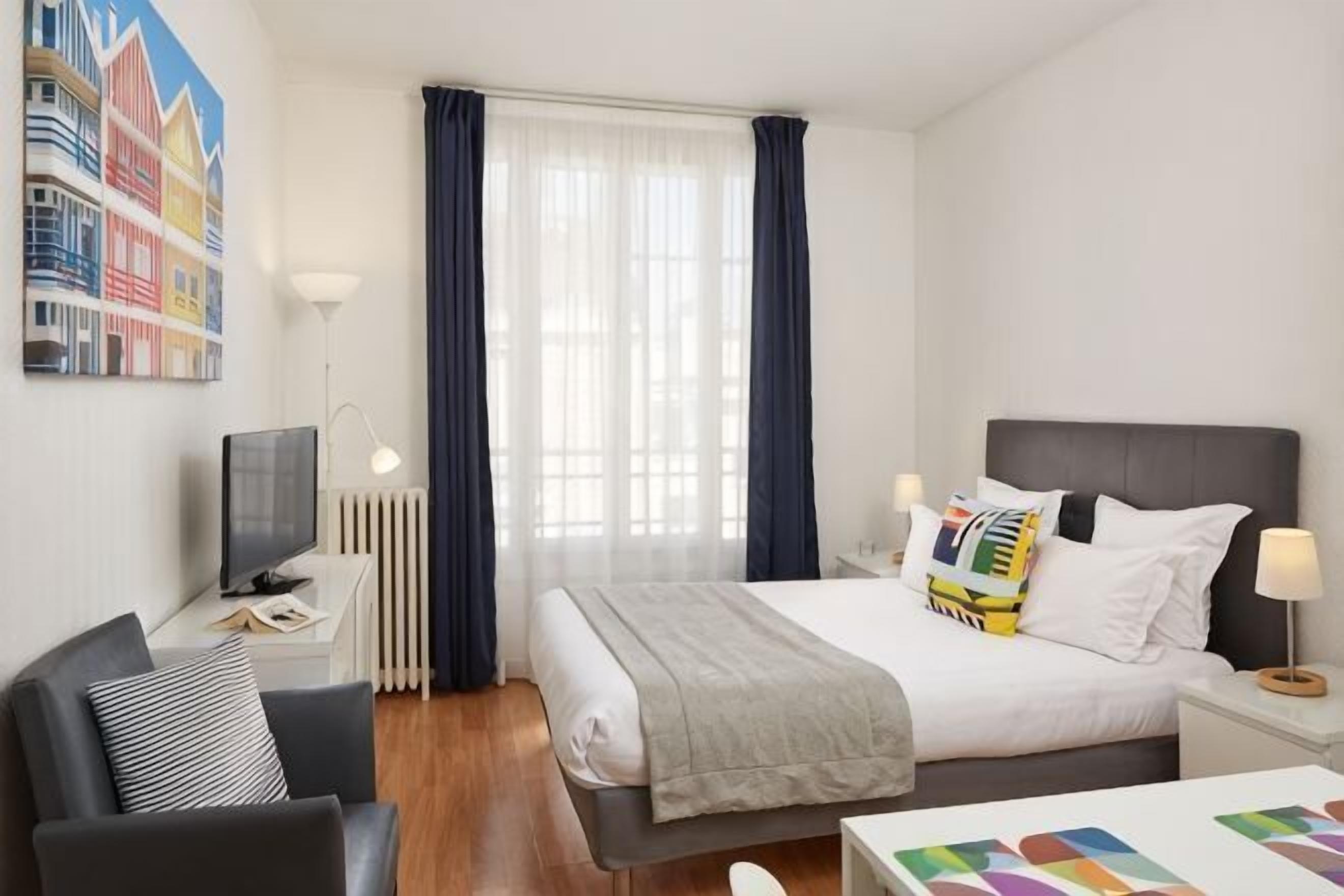 Une chambre d’hôtel comprenant un lit, des tables de chevet, une chaise, un bureau, une télévision et une fenêtre avec des rideaux.