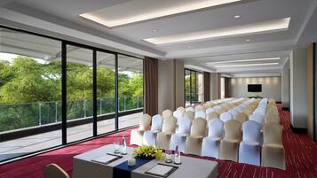 Un salón de conferencias con filas de sillas, una larga mesa y una alfombra roja, y ventanas amplias que dan a vistas de vegetación.