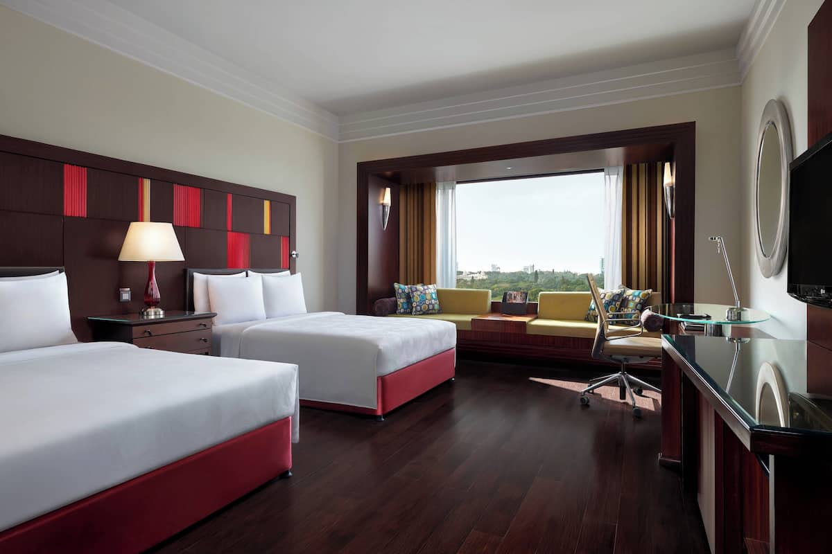 A hotel room with two beds, a desk, a chair, and a large window offering a city view.