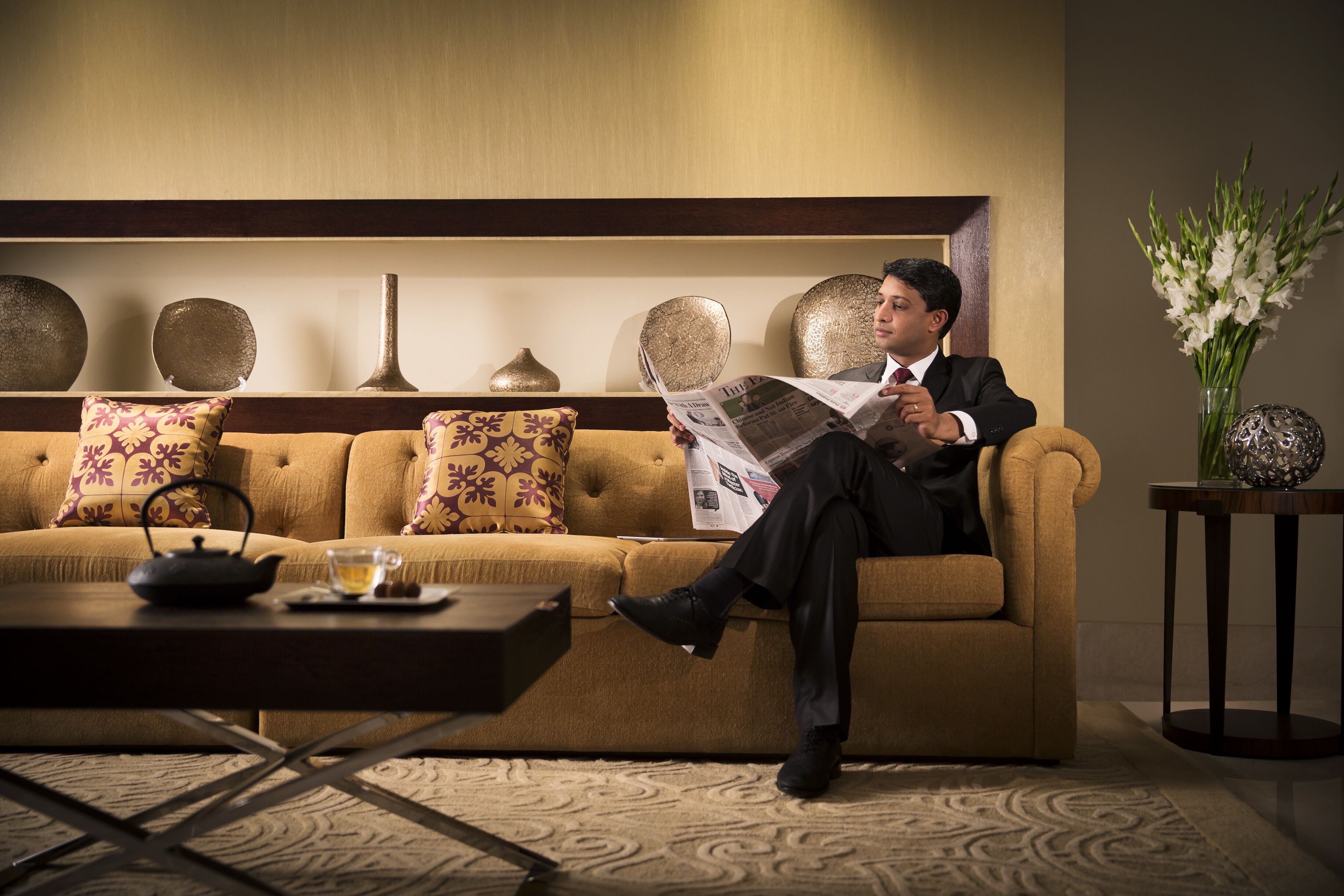 A man reading a newspaper on a sofa in a well-lit room with a decorative wall, a vase of flowers, and a small table with a teapot and cup.