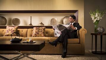 A man reading a newspaper on a sofa in a well-lit room with a decorative wall, a vase of flowers, and a small table with a teapot and cup.