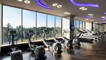 A gym with treadmills, elliptical machines, and exercise bikes, large windows with a view of greenery, and modern ceiling lighting.
