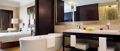 A modern hotel bathroom with a large mirror, a bathtub, two sinks, and a vanity with towels.