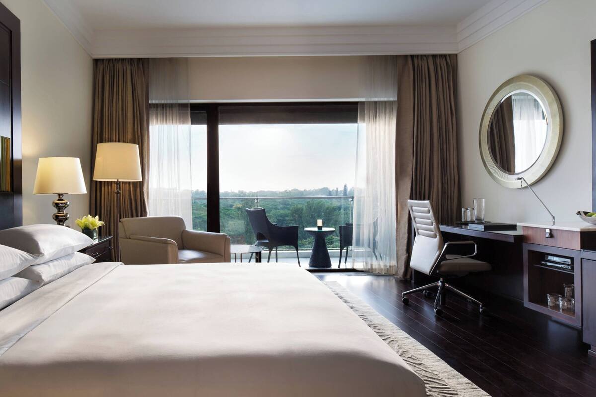 A hotel room with a large bed, a desk with a chair, a small table, and a view of the city through large windows.