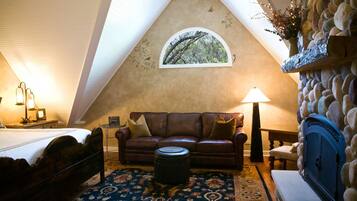 A cozy room with a sloped ceiling, a stone fireplace, a brown leather sofa, a round ottoman, and a bed with a white headboard.