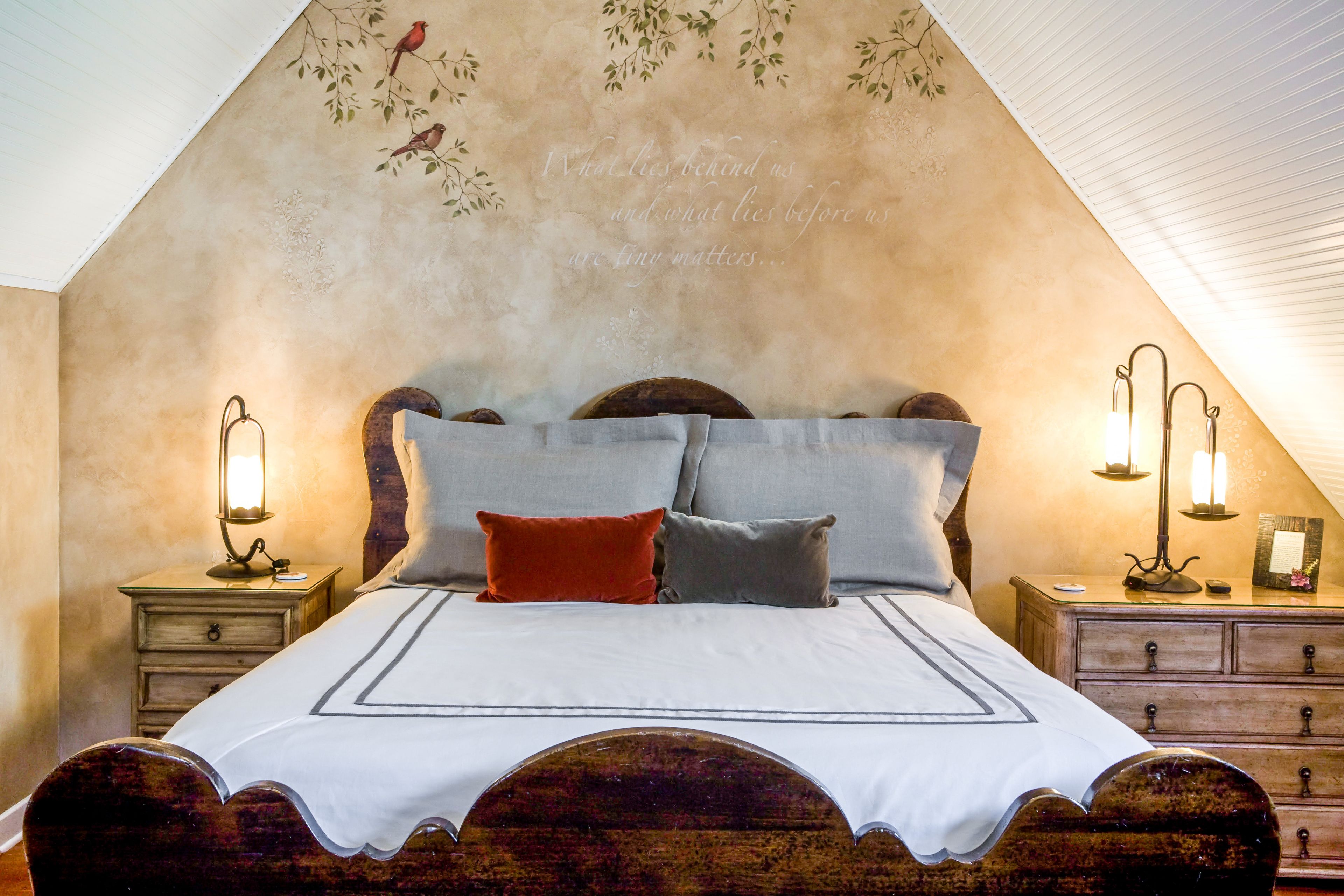 Une chambre à coucher avec un lit en bois, deux tables de chevet avec des lampes et un mur décoratif orné d’oiseaux et de motifs textuels.