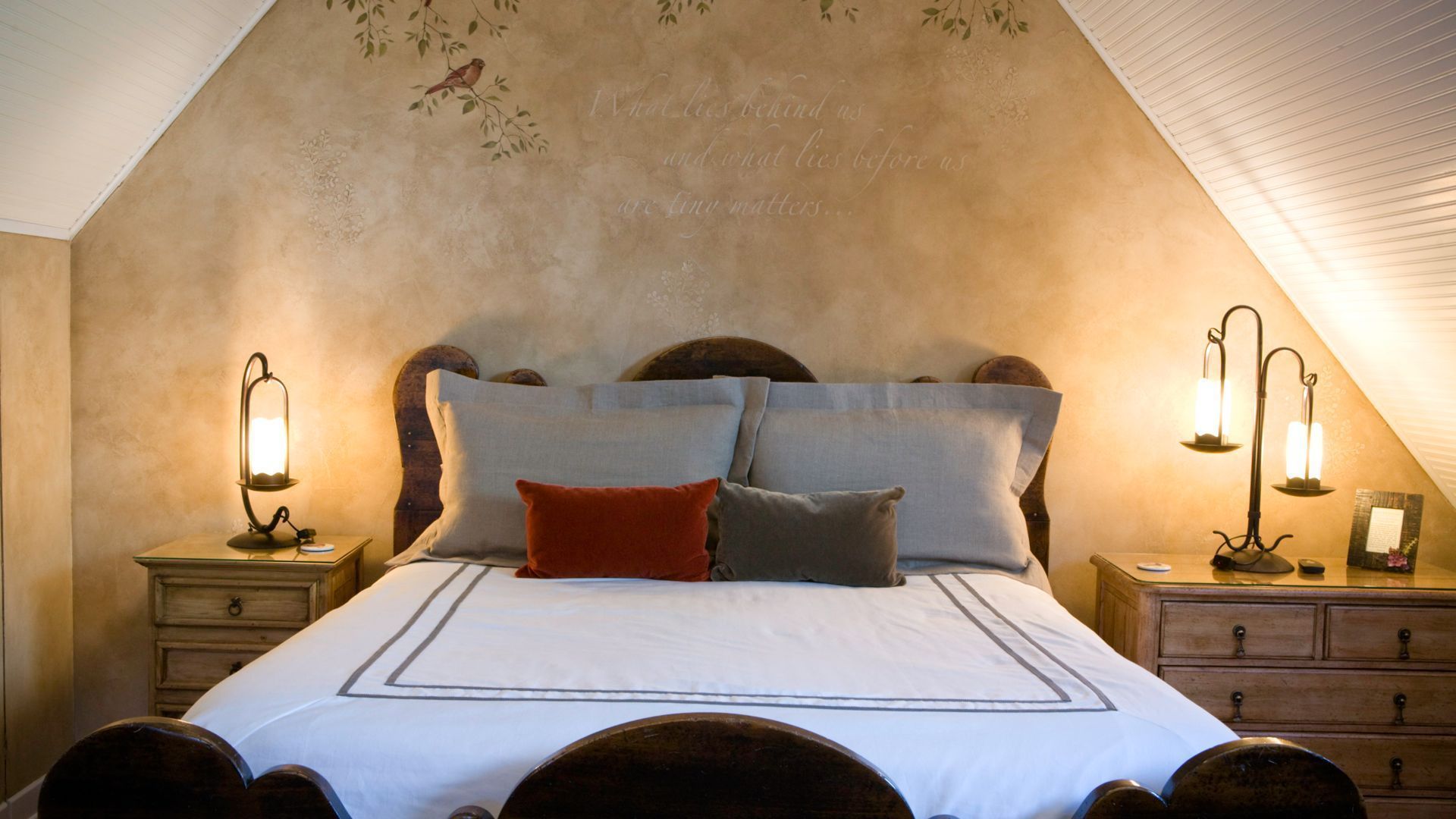 A cozy bedroom with a sloped ceiling, a large bed with multiple pillows, two bedside tables with lamps, and a decorative wall with a bird and floral pattern.