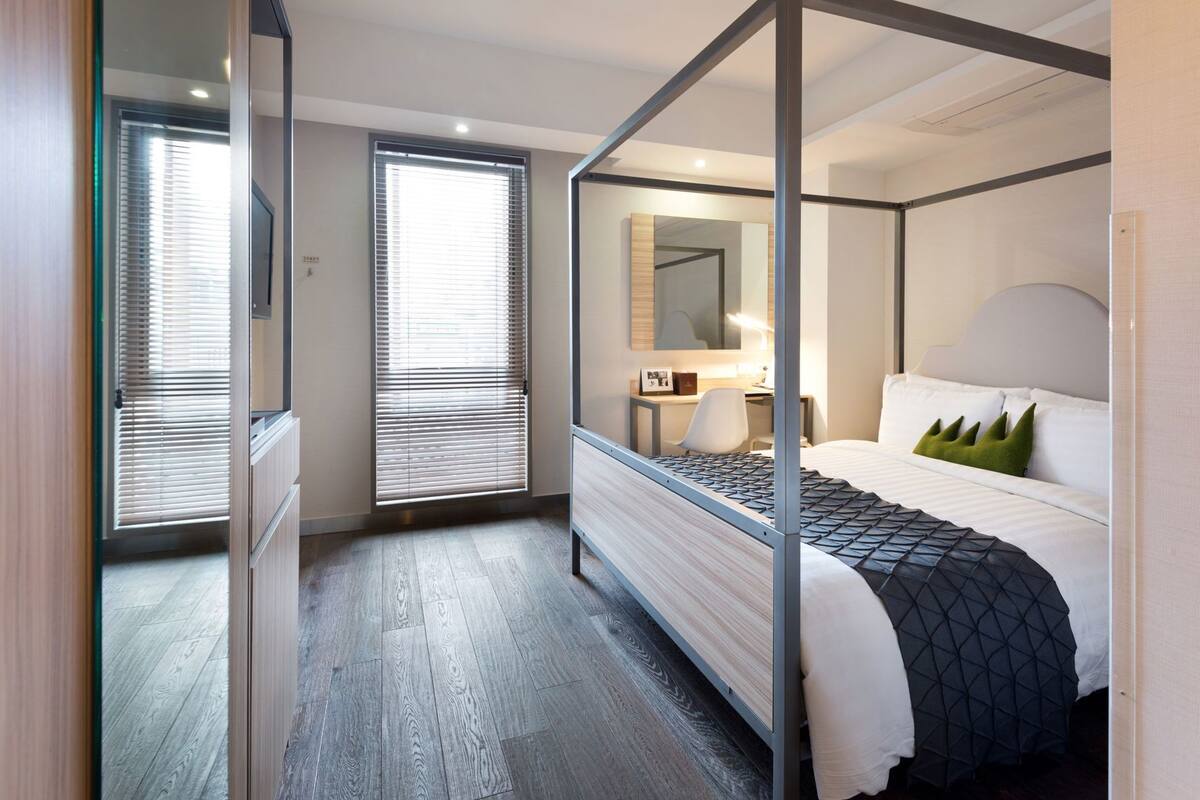 A modern hotel room with a four-poster bed, a desk with a chair, a large mirror, and a window with blinds.