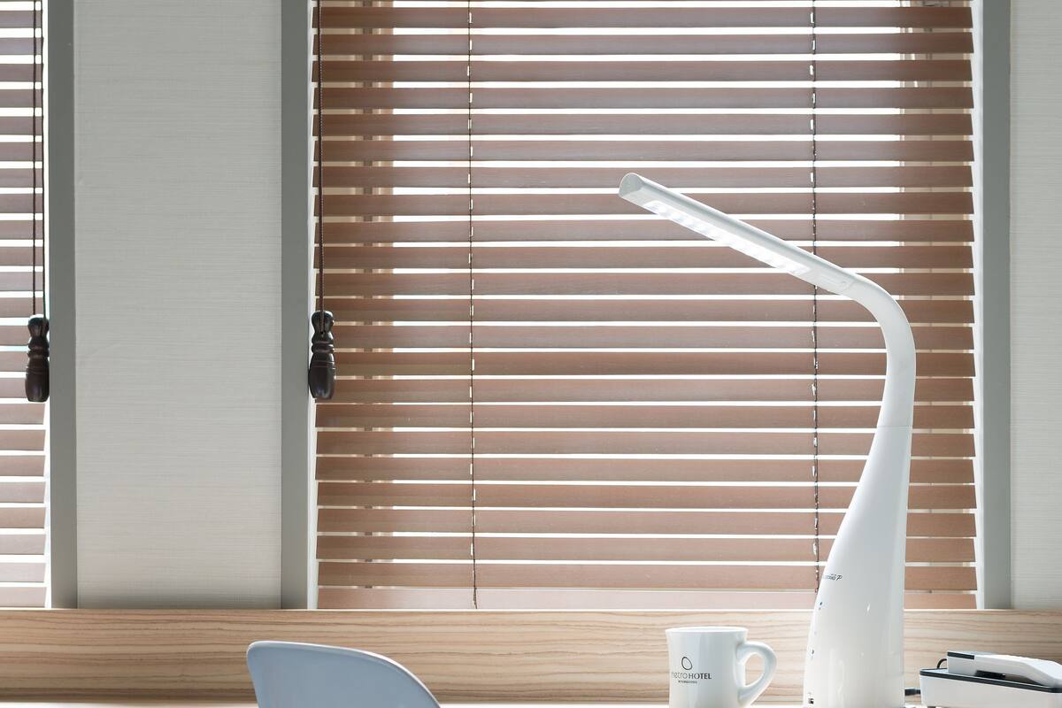 A modern bedroom with a bed, a white chair, a desk with a lamp, and a window with blinds.