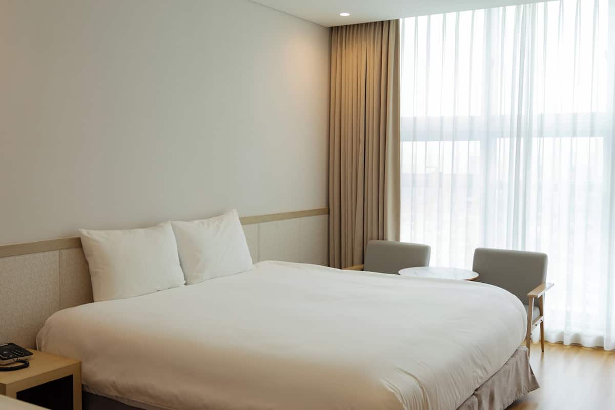 Ein Hotelzimmer mit einem großen Bett, einem Nachttisch mit Telefon, einem Sessel und einem Fenster mit Vorhängen.