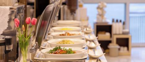 A buffet setup with various dishes, a vase of tulips, and a view of the sea in the background.