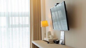 Habitación de hotel con televisor de pantalla plana montado en la pared, una mesita de noche con lámpara, cafetera y control remoto.