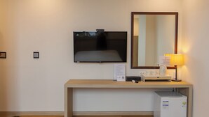 Una habitación de hotel con televisión de pantalla plana montada en la pared, una mesita de noche con lámpara, un refrigerador pequeño y un espejo sobre la mesa.