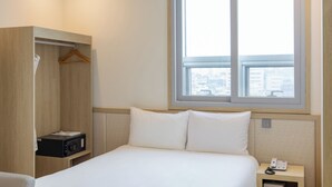 Habitación de hotel con cama, mesita de noche con teléfono, armario de madera y ventana con vista a la ciudad.
