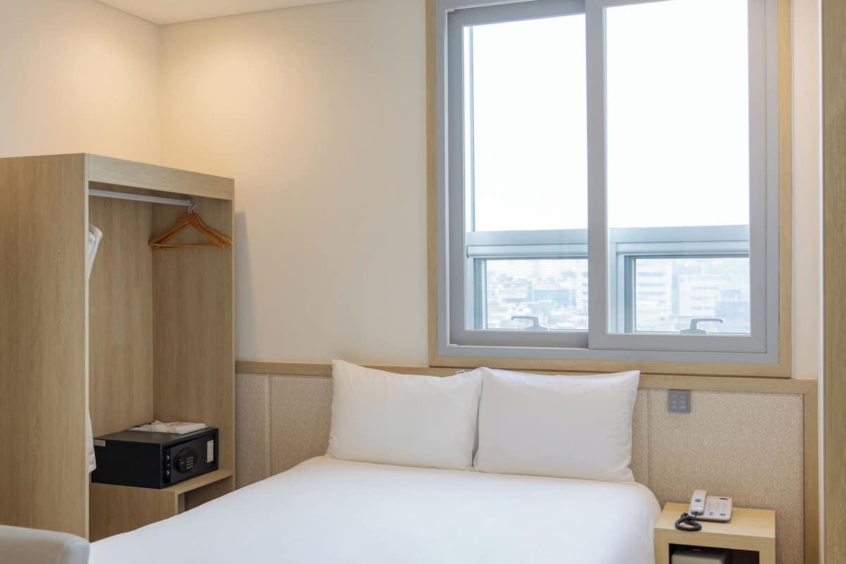 Ein Hotelzimmer mit einem Bett, einem Nachttisch mit Telefon, einem Holzschrank und einem Fenster mit Blick auf die Stadt.