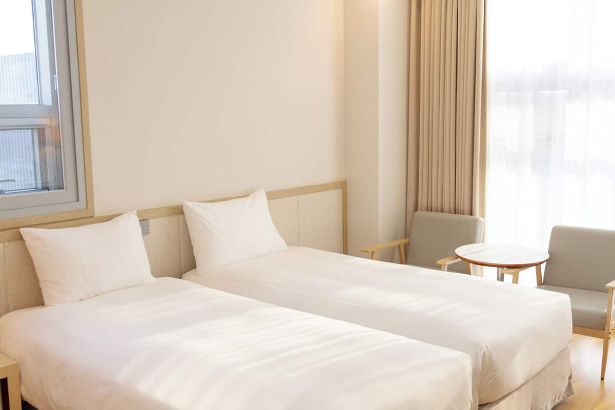 Ein Hotelzimmer mit zwei Betten, einem kleinen runden Tisch, einem Stuhl und einem Fenster mit Vorhängen.
