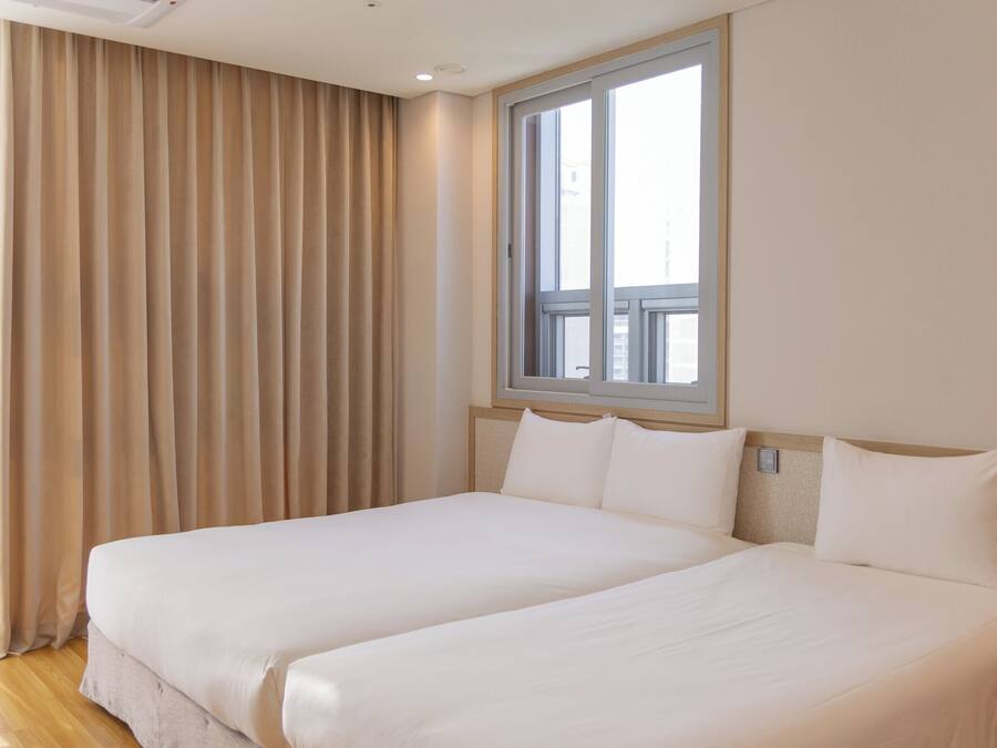 Una cama doble con sábanas blancas, una ventana con vistas y cortinas color beige.