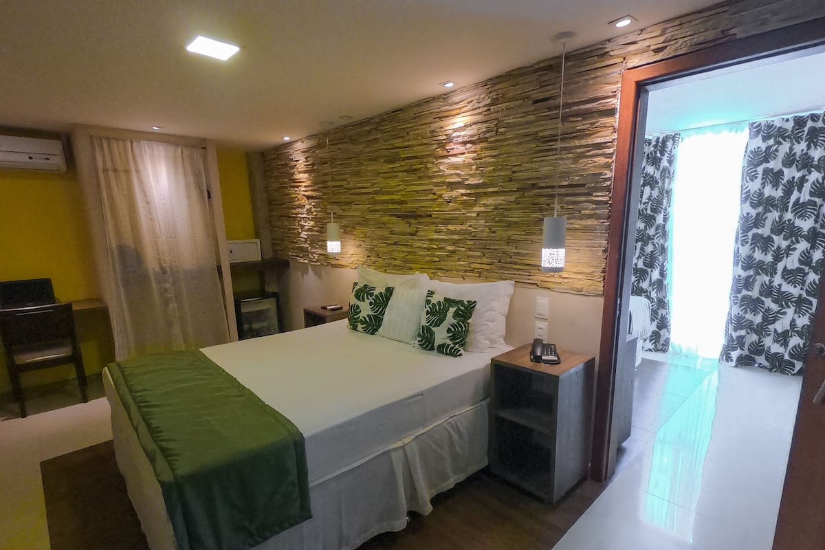 A hotel room with a bed, a desk, a chair, a nightstand, and a stone wall.