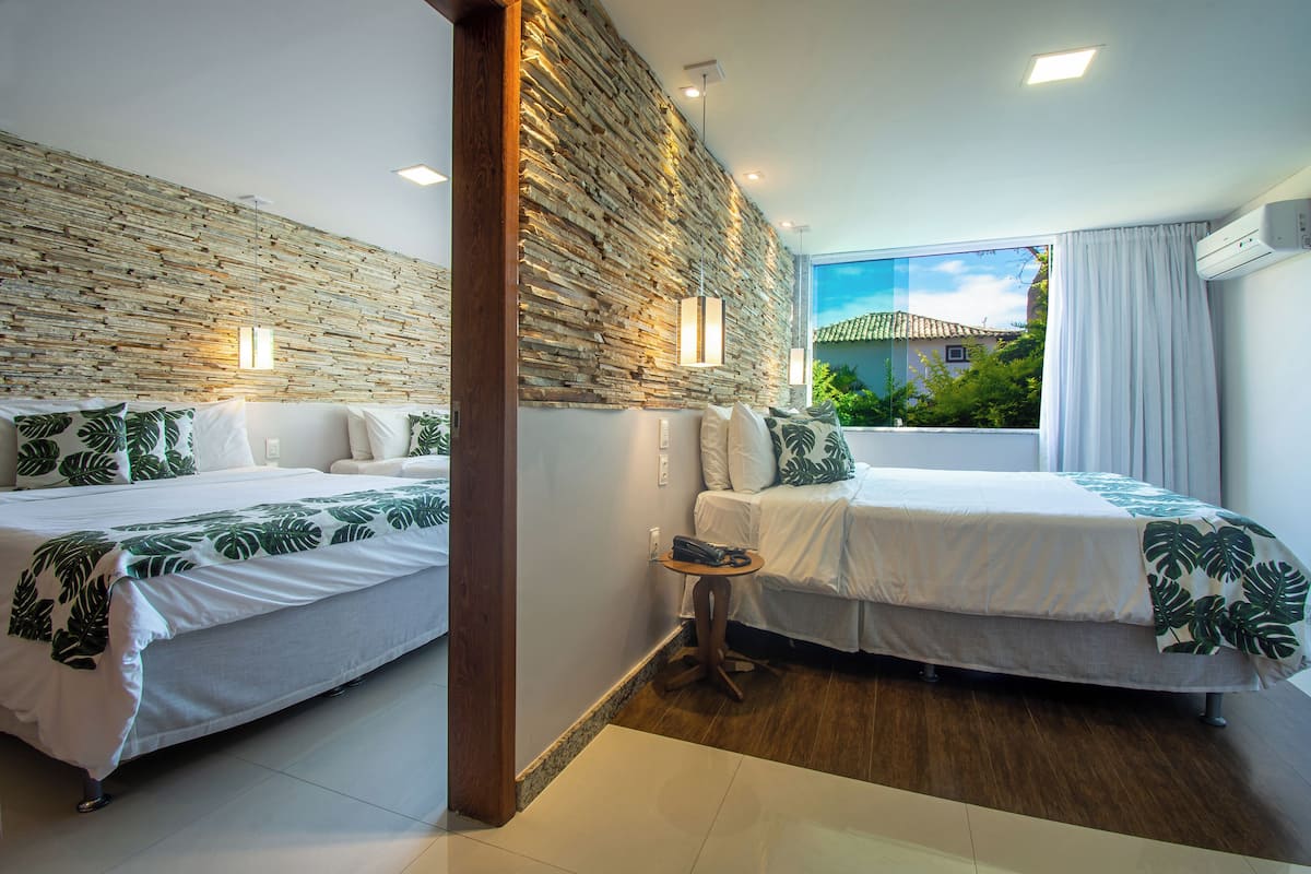 A hotel room with two beds, a stone wall, a small table, and a window with a view of greenery.