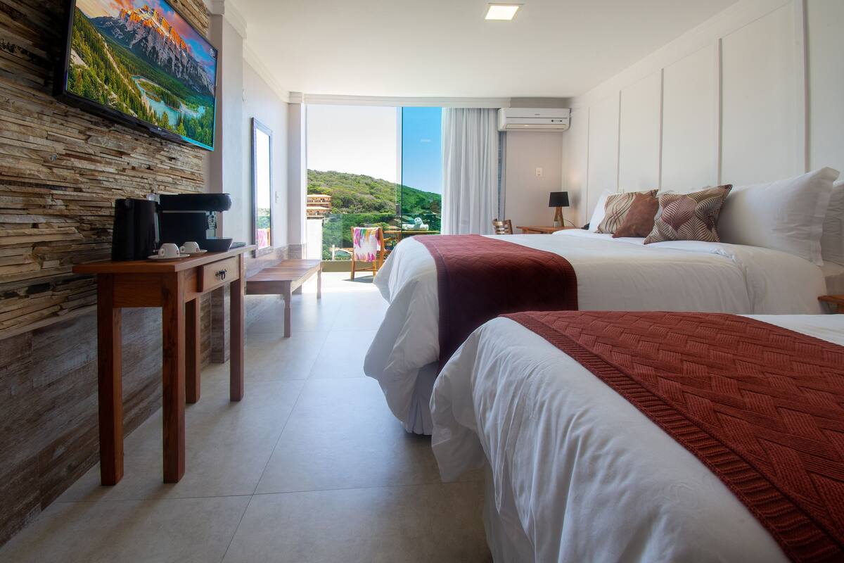 A hotel room with two beds, a TV mounted on a stone wall, a balcony with a scenic view, and a wooden desk with a coffee maker.