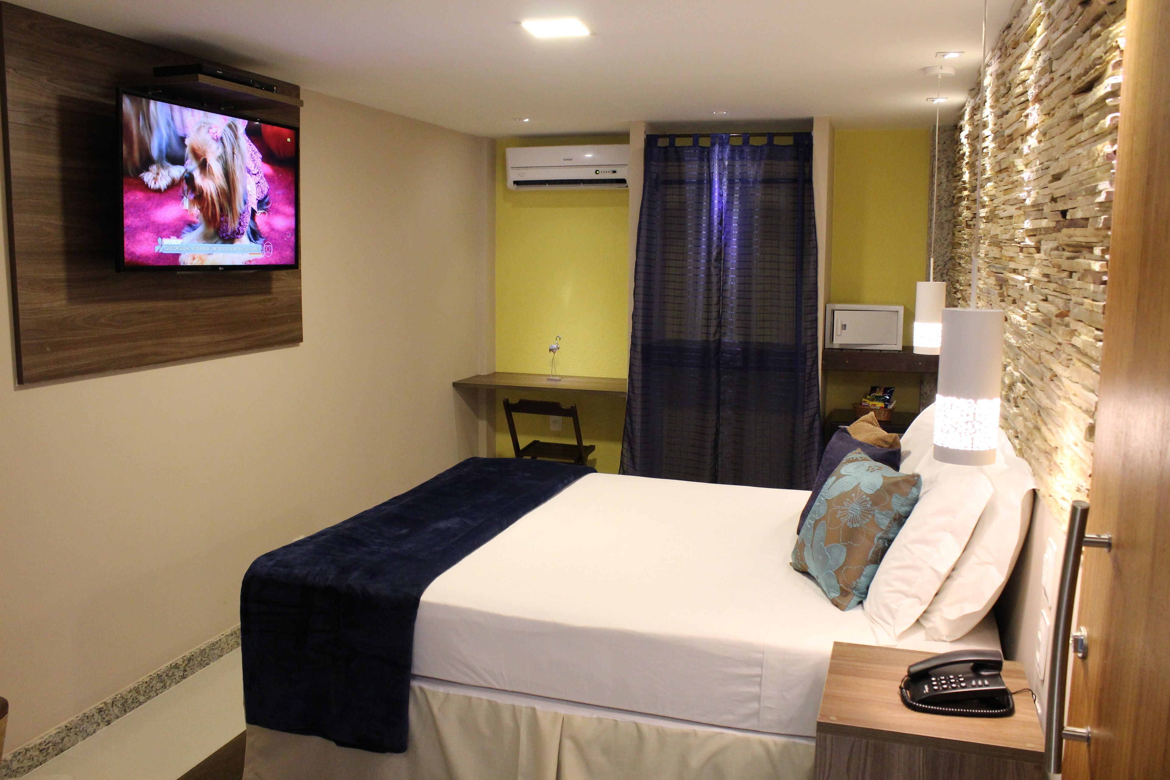 A hotel room with a bed, a television mounted on the wall, a desk with a chair, and a bedside table with a lamp and a telephone.