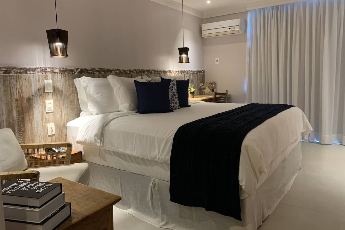A modern bedroom with a large bed, two pendant lights, a wooden headboard, a bedside table with books, and a window with curtains.