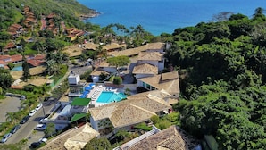 An aerial view of a coastal residential area with a swimming pool, houses with tiled roofs, and palm trees. - Hotel Experience João Fernandes (Búzios)