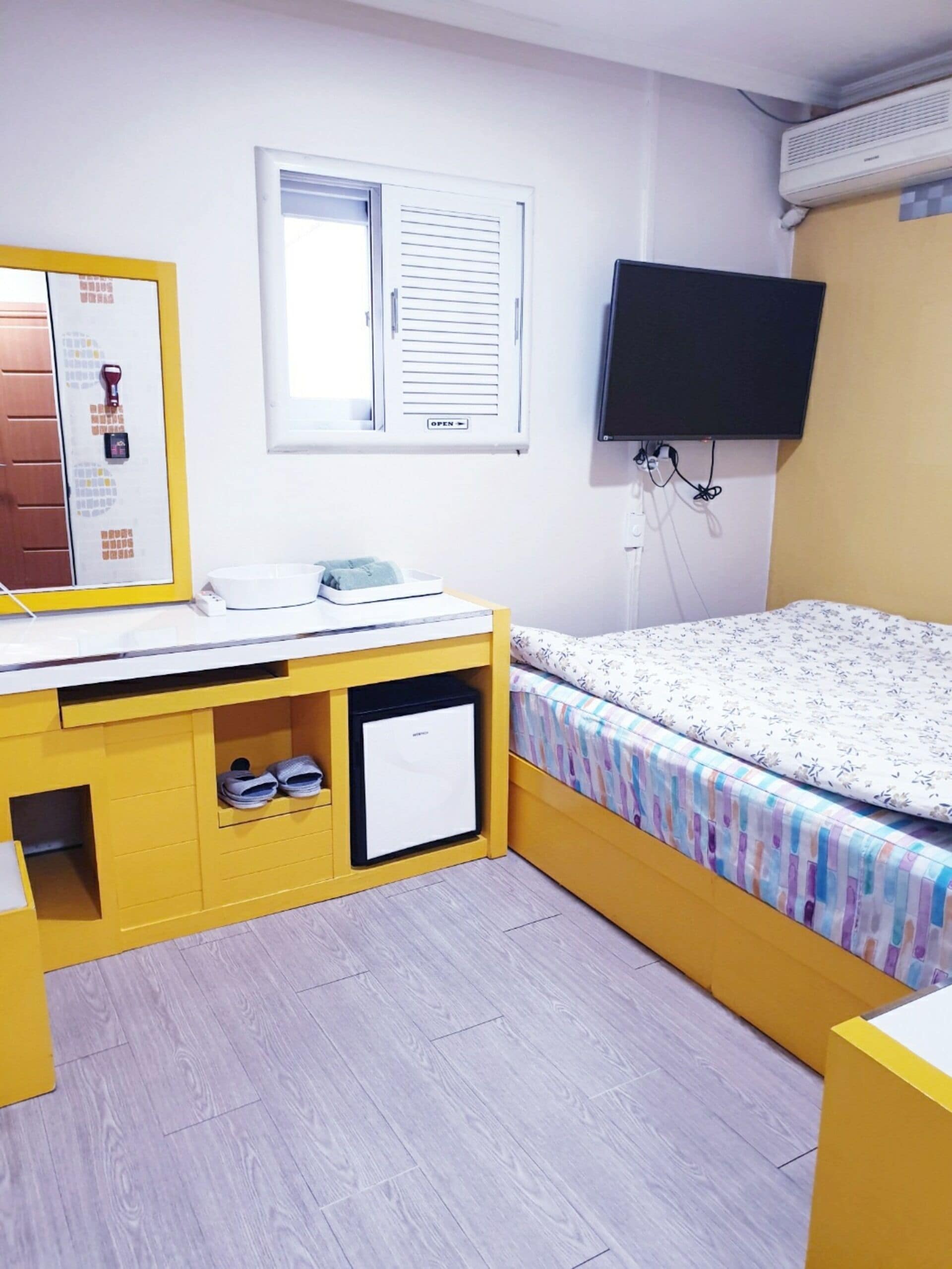 Une chambre compacte comprenant un lit, un bureau avec un miroir, un petit réfrigérateur et un téléviseur fixé au mur.