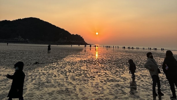 Une scène de plage avec des personnes qui se promènent et qui sont debout, un coucher de soleil en arrière-plan et une colline au loin.