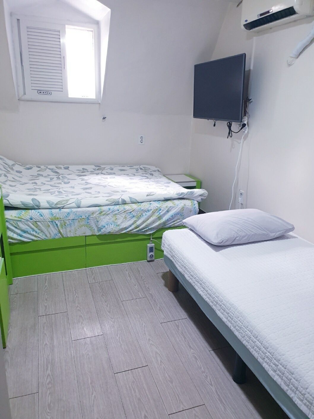 A small, well-lit bedroom with a single bed, a green nightstand, a wall-mounted TV, and a window with blinds.