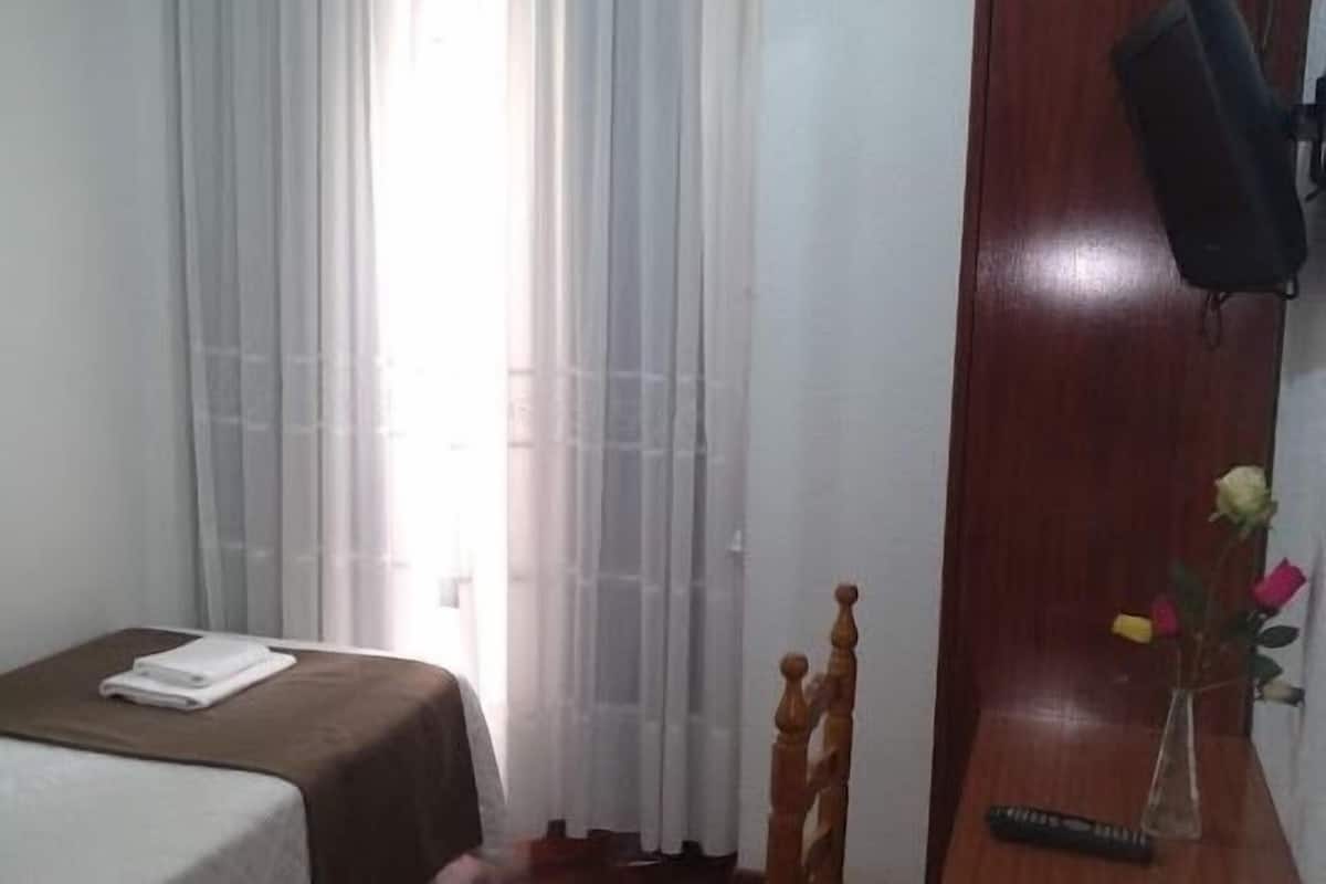 A hotel room with a bed, a wooden chair, a vase with flowers, a remote control, and a wooden door.