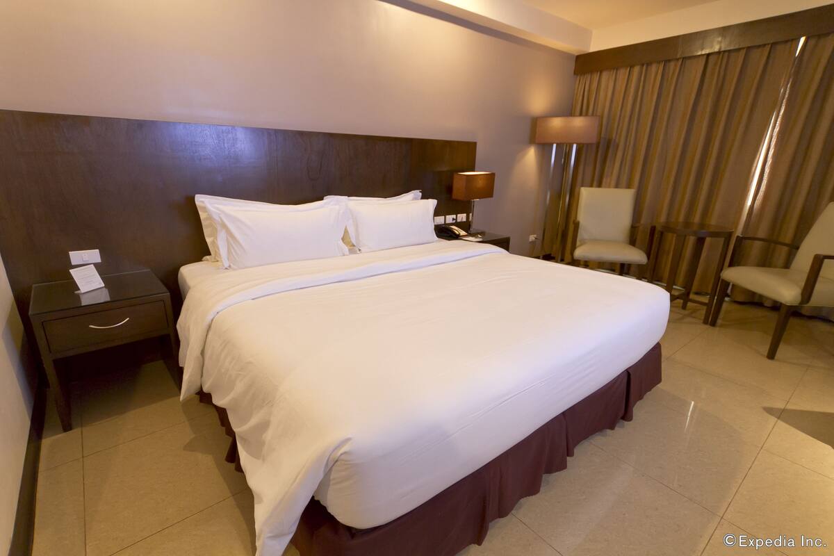 Una habitación de hotel con una cama grande, una mesita de noche con lámpara, una silla y una cortina.