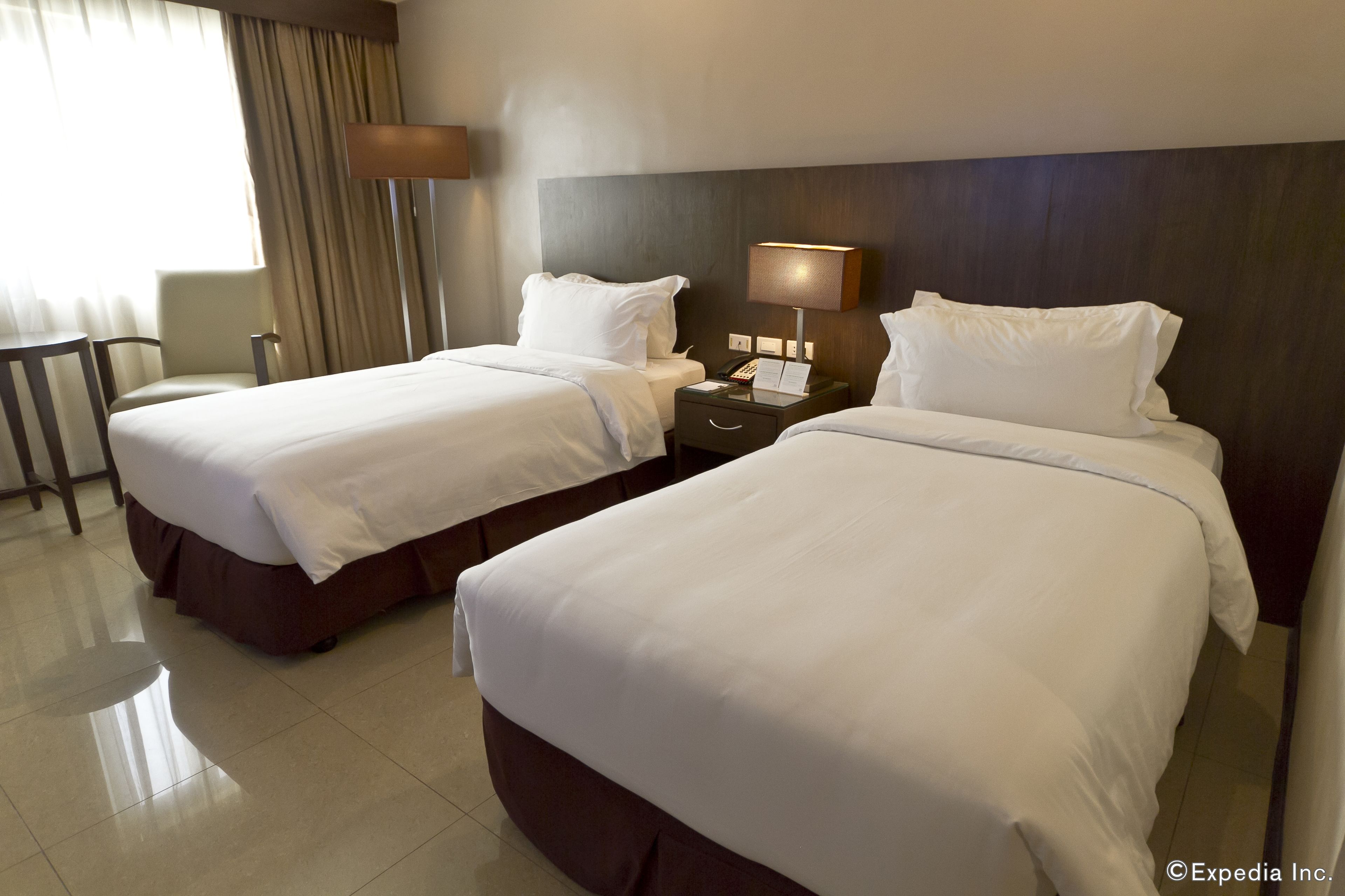 A hotel room with two beds, a bedside table with a lamp, a chair, and a desk.