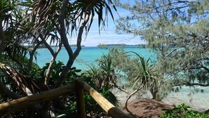 Eine Holzterrasse führt zu einem Strand mit klarem, türkisblauem Wasser und fernen Inseln.